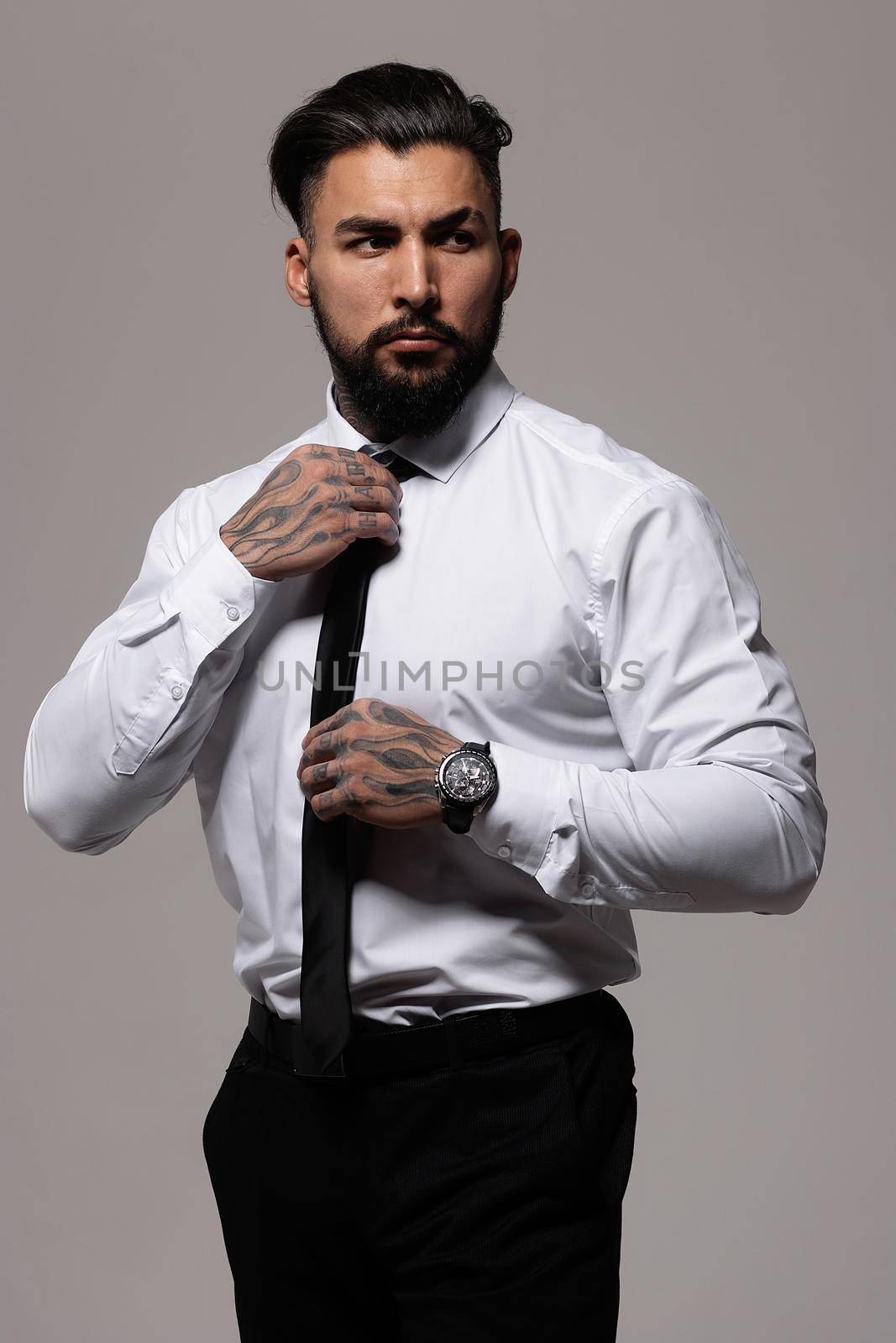 Confident Hispanic bearded man with tattooed hands in white shirt and black trousers adjusting tie and looking away while standing against gray background