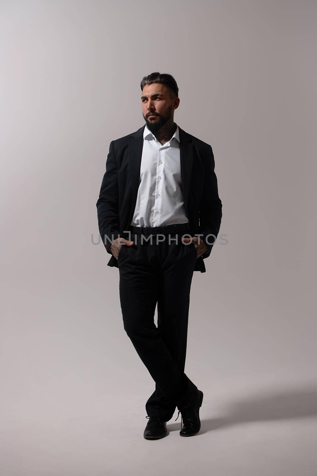 Bearded Hispanic guy in dark vest and white shirt with tie looking at camera with hands in pockets in studio