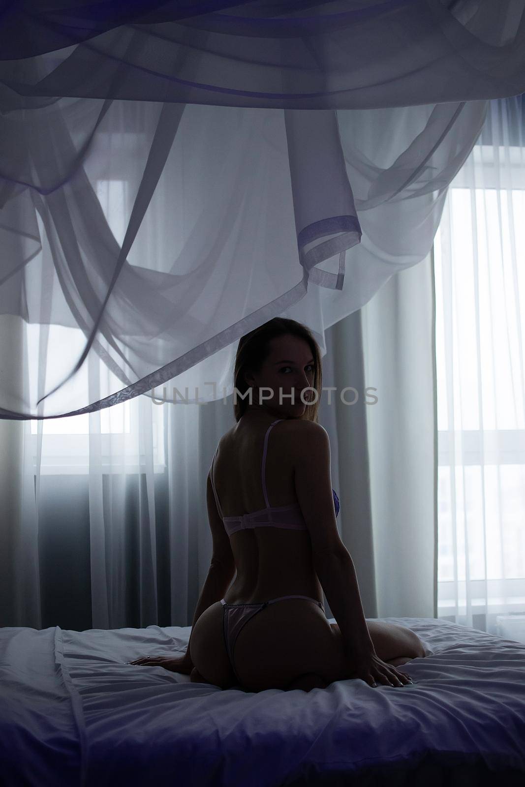 Seductive slim female in purple lingerie sitting on soft bed in room and looking up
