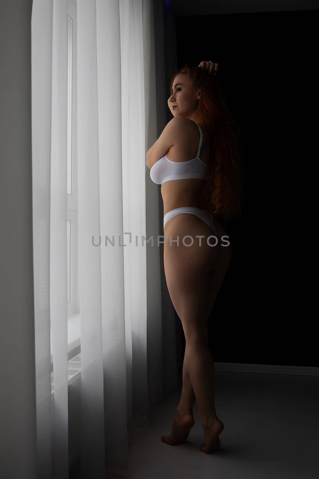 Slender sexy female in white lace underwear touching long hair while standing with closed eyes near window in room
