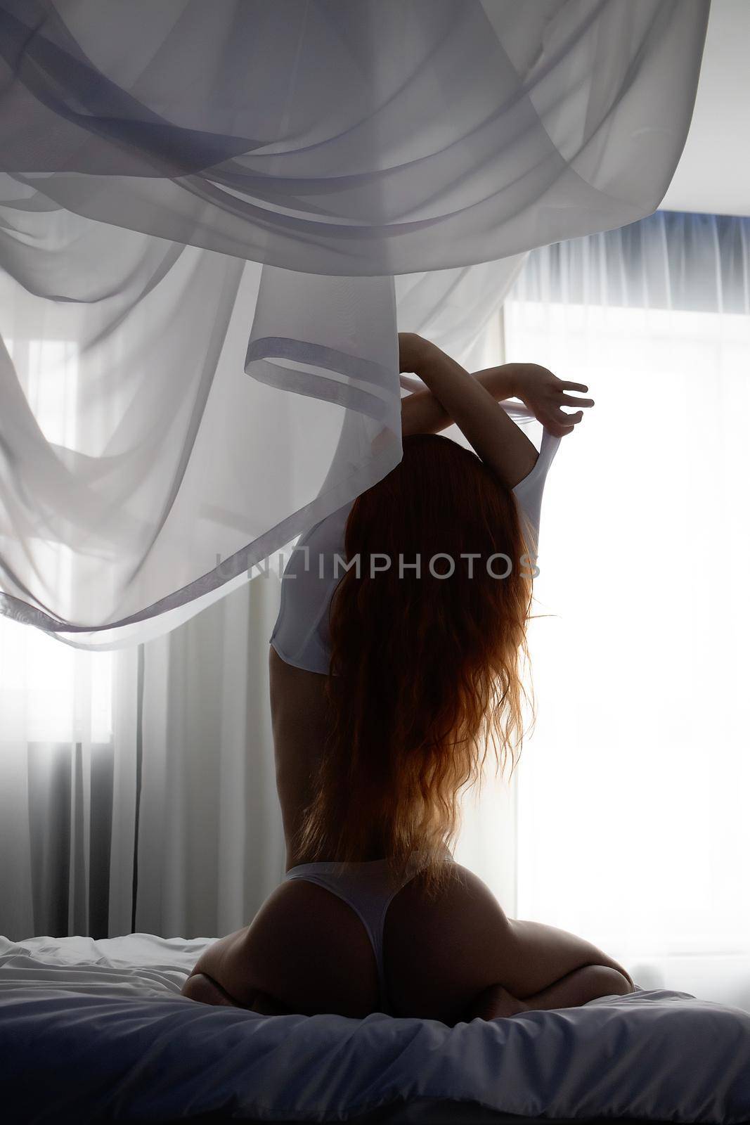 Back view of sexy female with long red hair taking off bra while sitting on soft bed at home