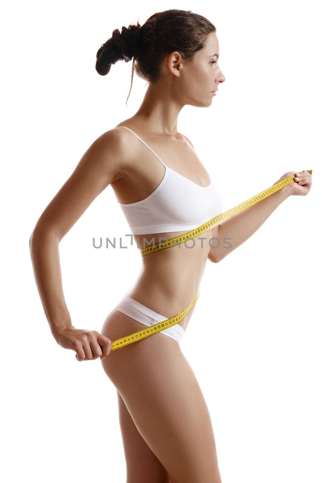 Perfect, slim, toned, young body of an alluring lady in white underwear measuring herself with a measuring tape while posing sideways isolated on white. Beauty treatment. Plastic surgery and aesthetic cosmetology concept. Augmentation or reduce of a breast, cellulite removal. High resolution, close-up shot.