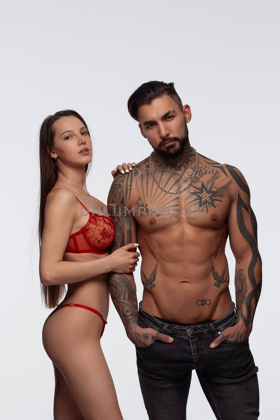 Confident tattooed bearded male standing with seductive female partner wearing red lace lingerie in studio and looking at camera
