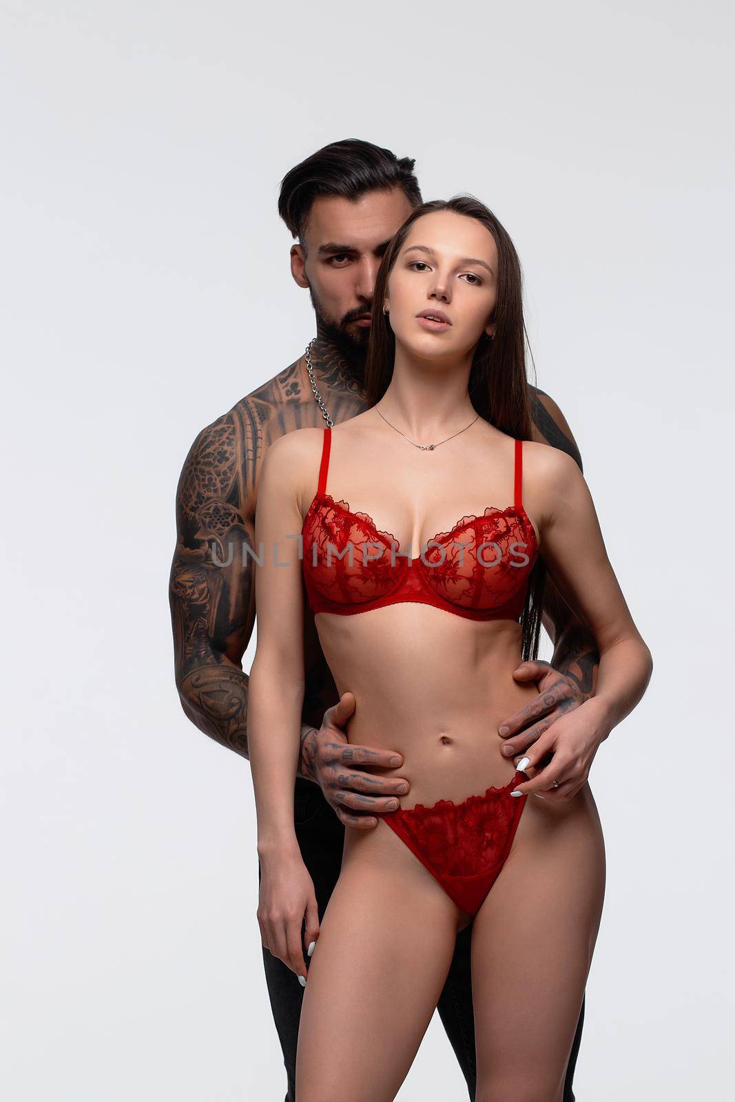 Confident tattooed bearded male standing with seductive female partner wearing red lace lingerie in studio and looking at camera
