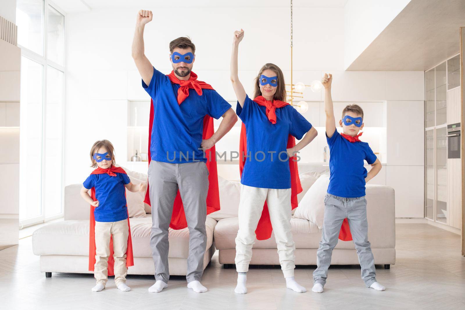 Full body of cheerful parents with two children in casual clothes and red superhero cloaks playing heroes at home