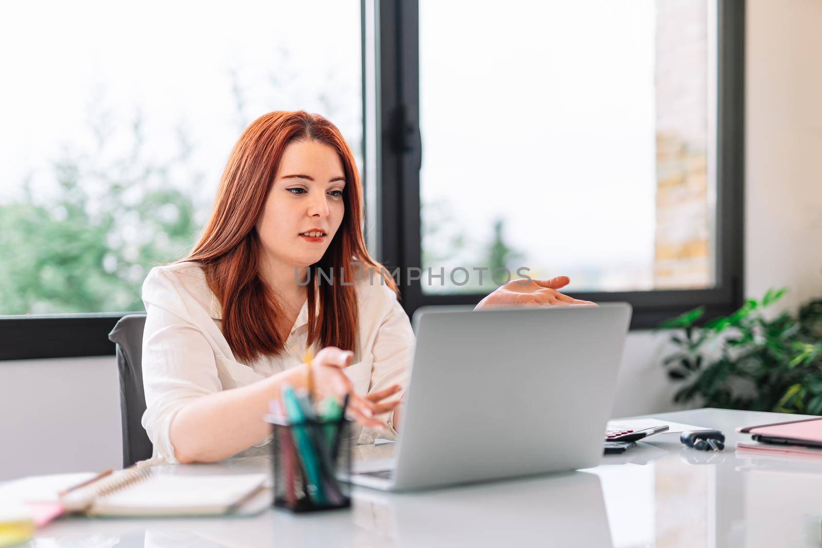 Pretty young redheaded pretty woman teleworking from home in white shirt. She is making a work video call explaining something. Very bright environment with a large window behind and natural light. Young businesswoman and entrepreneur working on white laptop.