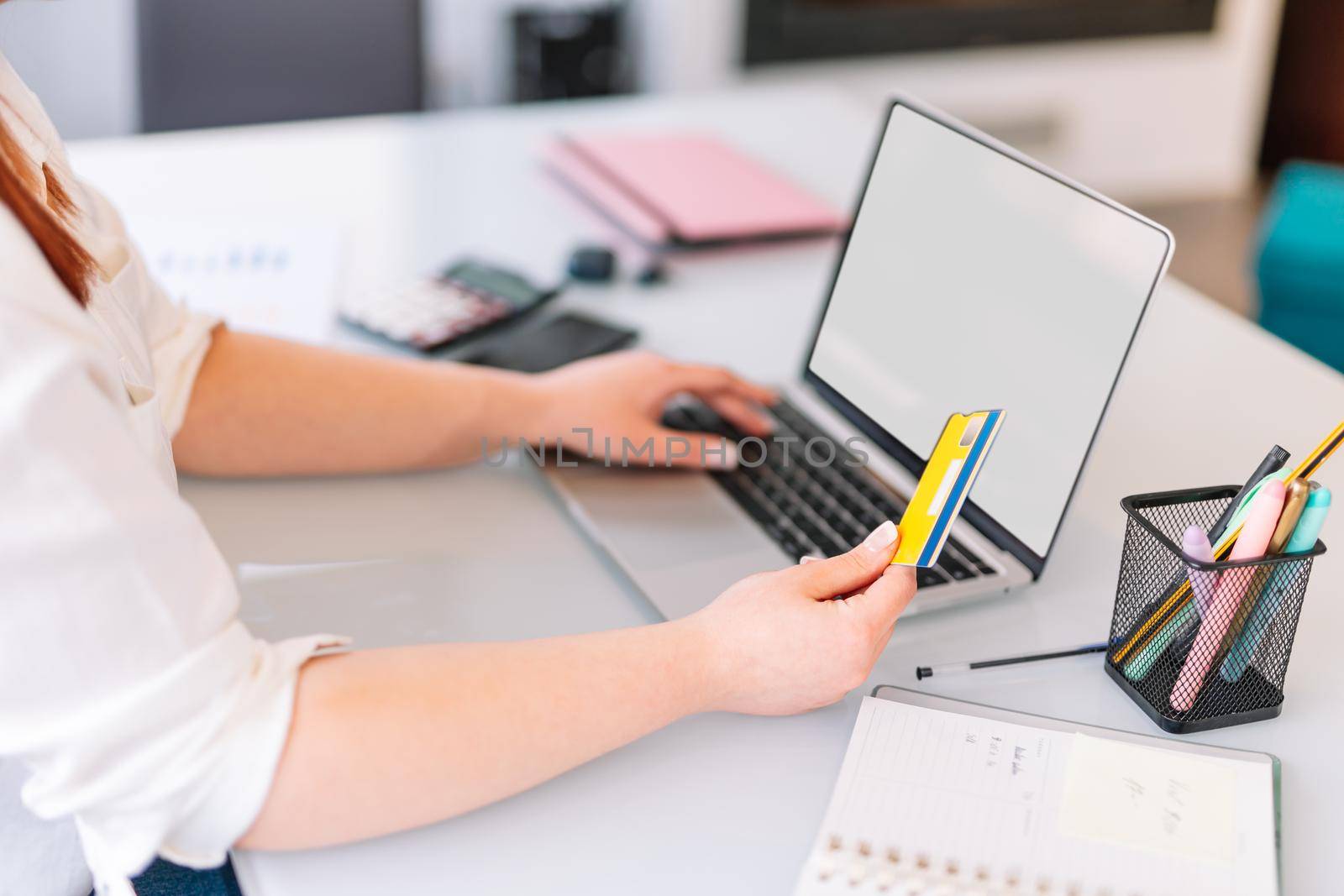 Pretty young redheaded girl using her debit or credit card to shop online. She is using the laptop and her bank card to pay and shop online. She is at her desk working in the office or at home. Blank laptop screen