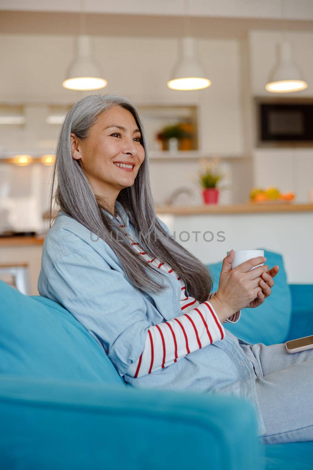 Joyful female person looking away and smiling while resting on sofa and holding mug of hot drink