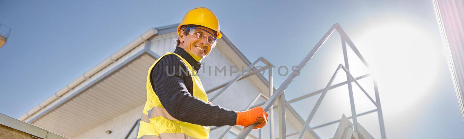 Joyful matured man in safety helmet looking at camera and smiling while walking up staircase at industrial plant