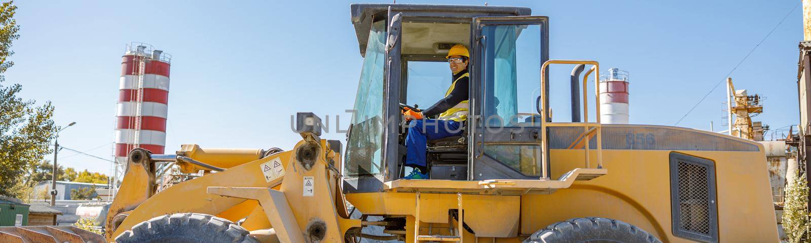 Joyful male worker looking at camera and smiling while sitting in tractor driver cabin at industrial plant