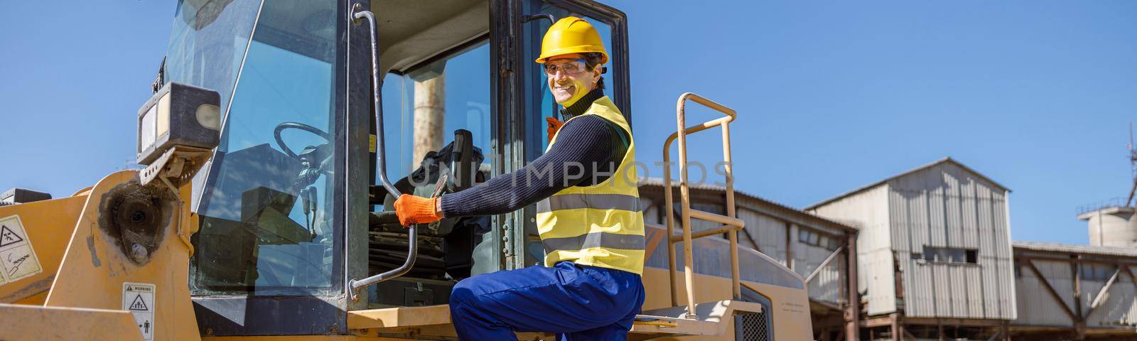 Cheerful male engineer standing on tractor stairs outdoors by Yaroslav_astakhov