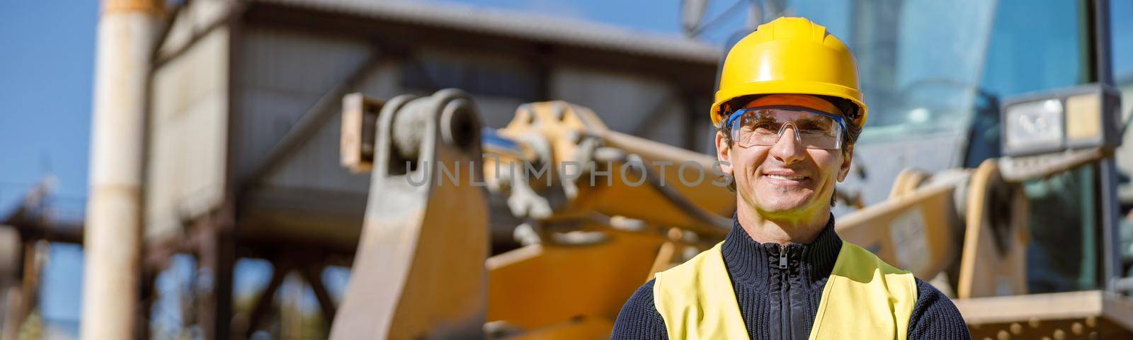 Joyful matured man engineer wearing safety helmet, glasses and vest while keeping arms crossed and smiling