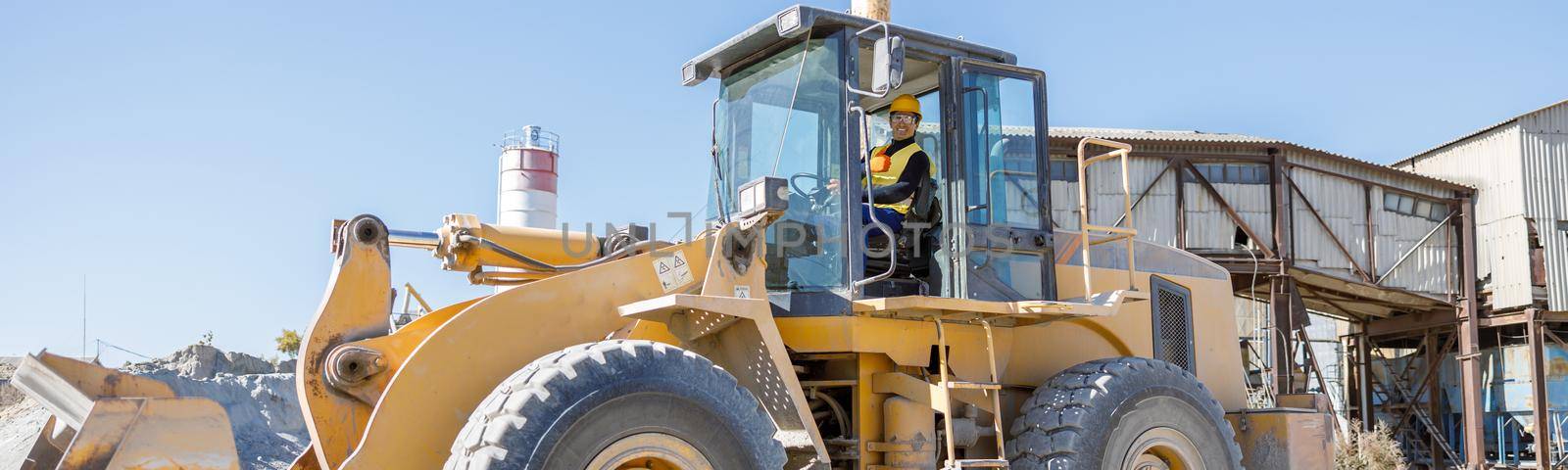 Cheerful male worker looking at camera and smiling while sitting in tractor driver cabin at industrial plant under blue sky