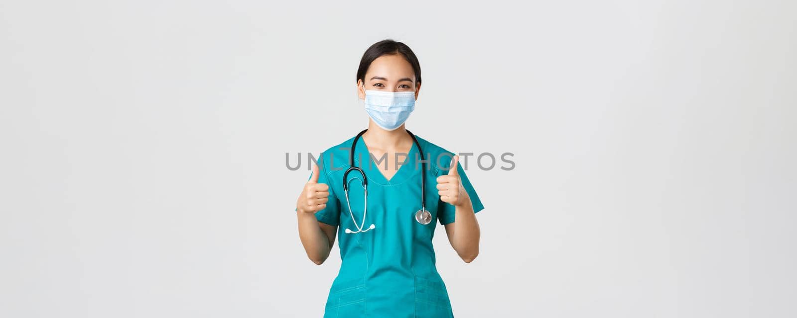 Covid-19, coronavirus disease, healthcare workers concept. Confident, smiling asian doctor, female nurse in scrubs and medical mask profive good quality service, show thumbs-up in approval by Benzoix