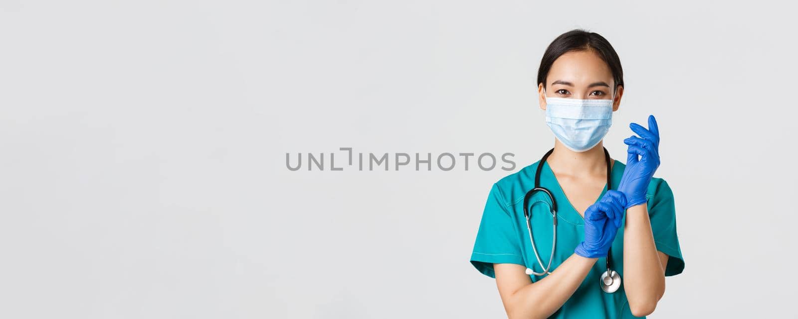 Covid-19, coronavirus disease, healthcare workers concept. Professional smiling asian female physician, doctor in medical mask and scrubs, put on rubber gloves ro examination, white background by Benzoix