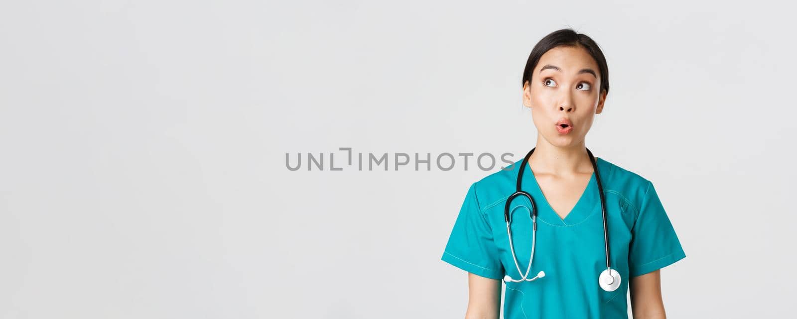 Healthcare workers, preventing virus, quarantine campaign concept. Impressed and amazed asian female doctor, nurse in medical scrubs looking upper left corner pleased, white background.