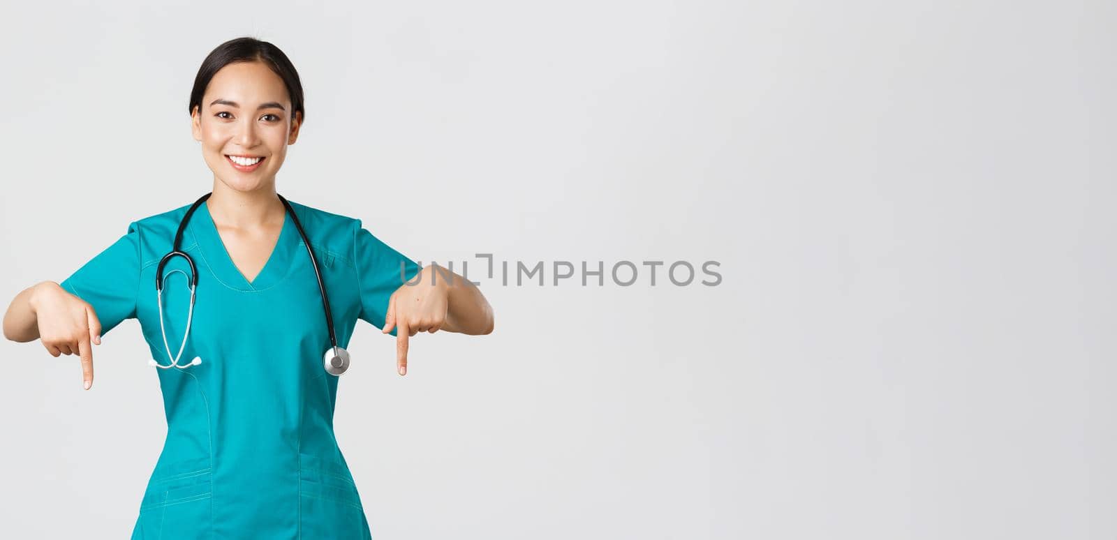 Covid-19, healthcare workers, pandemic concept. Smiling pleasant asian female doctor, therapist or physician in scrubs with stethoscope, pointing fingers down, show clinic banner.