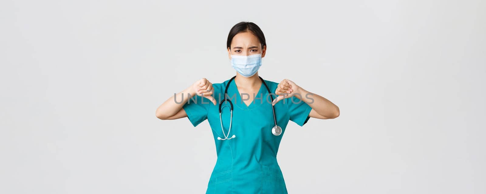 Covid-19, coronavirus disease, healthcare workers concept. Disappointed and upset asian female doctor in scrubs and medical mask showing thumbs-up, disagree or prohibit something, give advice by Benzoix