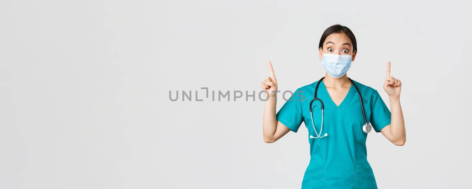 Covid-19, coronavirus disease, healthcare workers concept. Intrigued asian female doctor, physician or intern in medical mask and scrubs, looking curious, pointing fingers up, white background by Benzoix