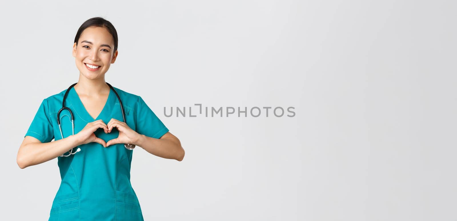 Covid-19, healthcare workers, pandemic concept. Lovely caring asian doctor, female nurse in scrubs showing heart gesture and smiling, taking care of patients with love, white background by Benzoix