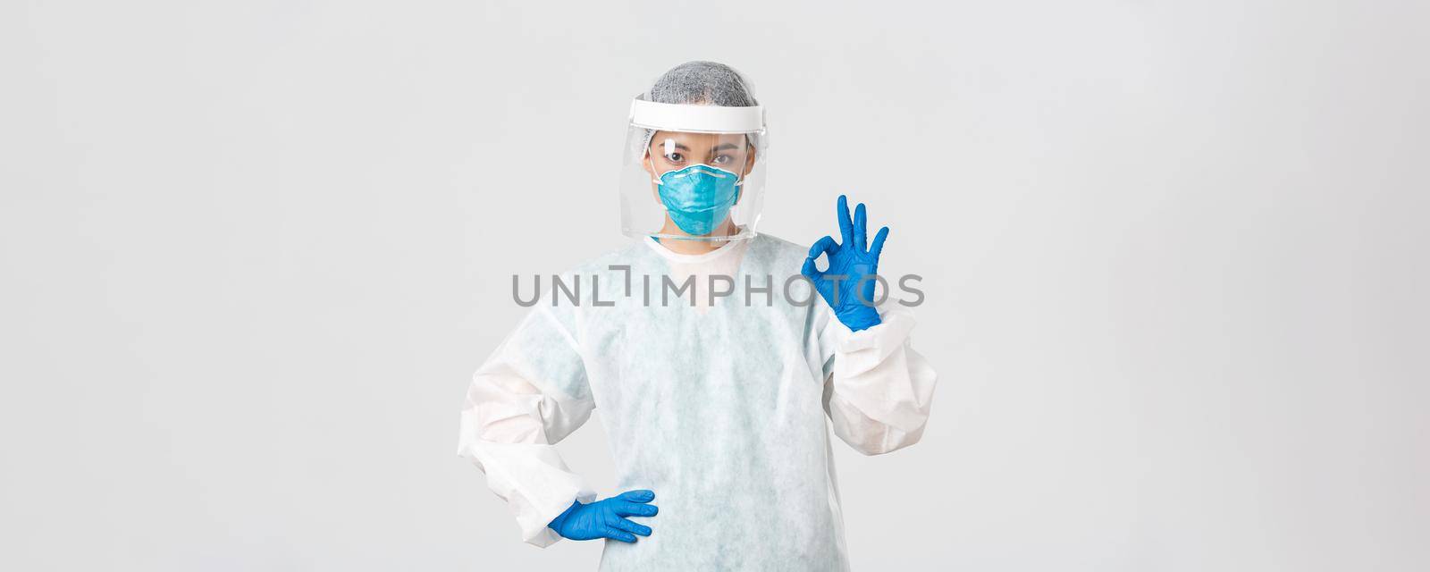 Covid-19, coronavirus disease, healthcare workers concept. Serious-looking professional female asian doctor in personal protective equipment, showing okay gesture, ensure safety of patient by Benzoix