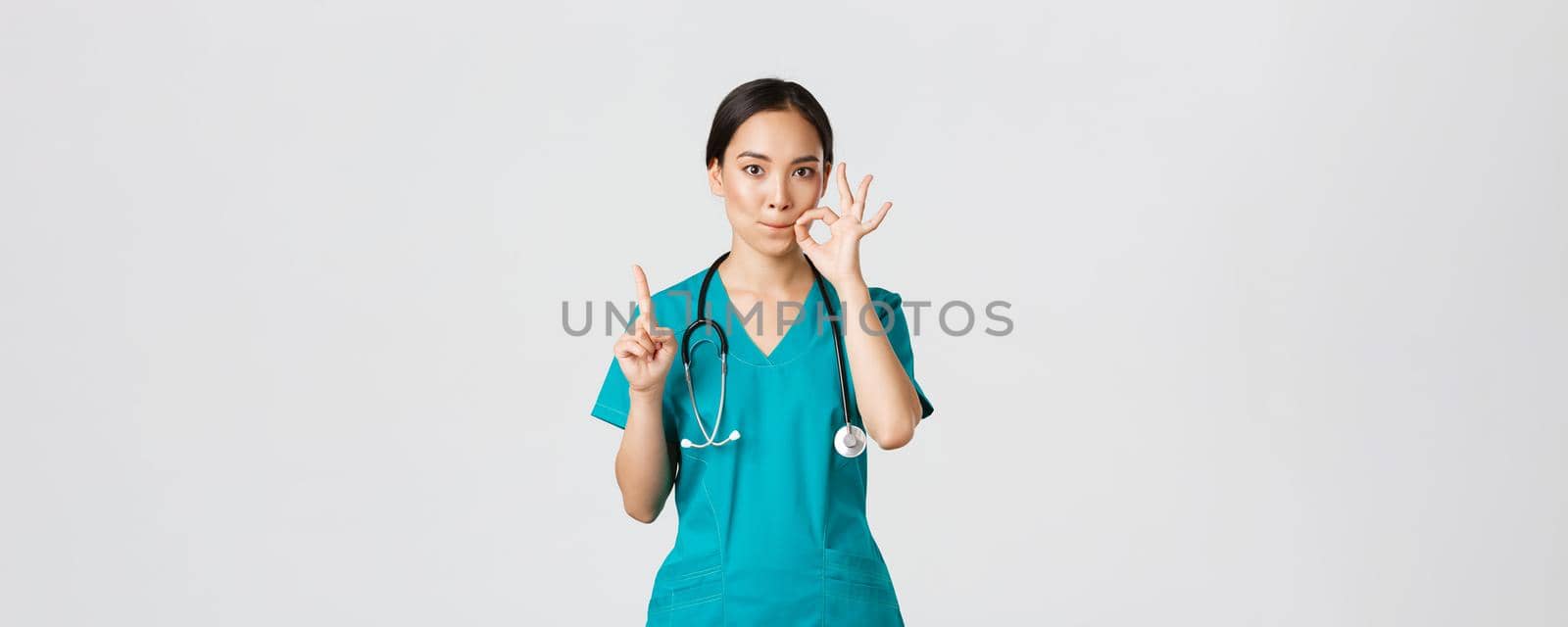 Covid-19, healthcare workers, pandemic concept. Serious-looking worried asian female nurse, physician asking keep secret, shaking finger and showing mouth seal, zipping lips gesture.