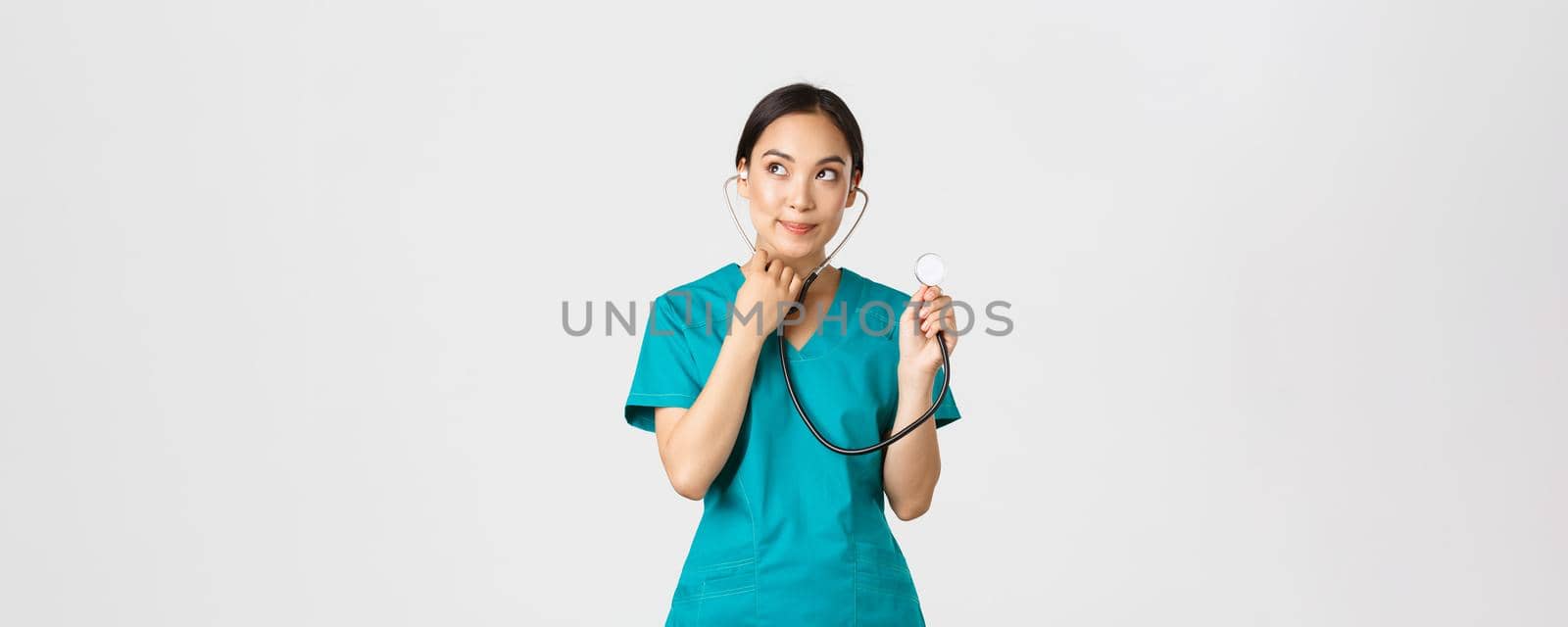 Covid-19, healthcare workers and preventing virus concept. Cute smiling asian female intern learn how listen lungs with stethscope, doctor examine patient, looking thoughtful upper left corner.