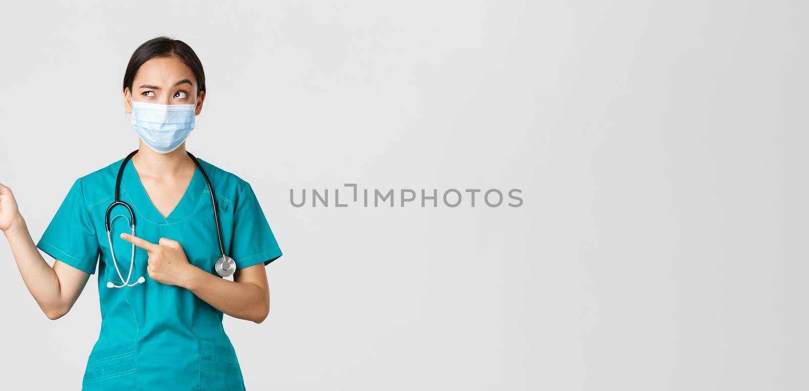 Covid-19, coronavirus disease, healthcare workers concept. Suspicious or doubtful asian female doctor in scrubs and medical mask, looking uncertain and pointing upper left corner, white background.