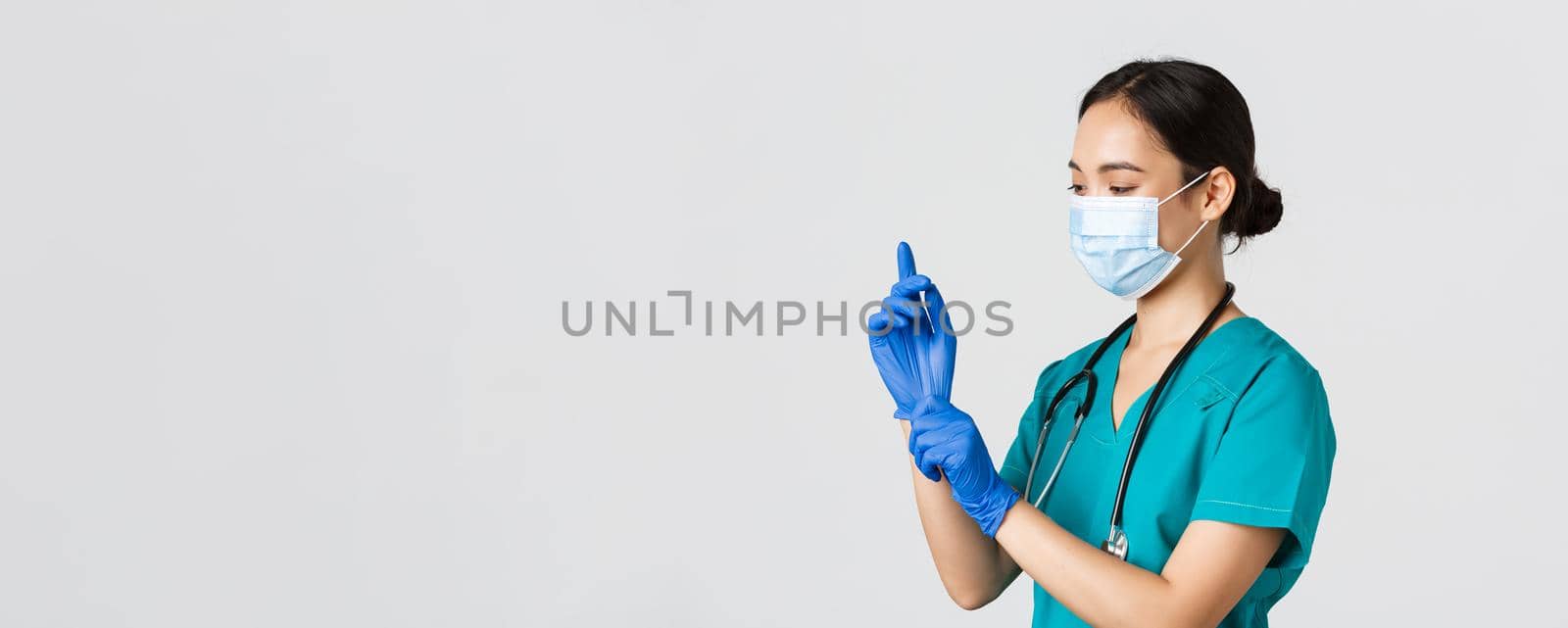 Covid-19, coronavirus disease, healthcare workers concept. Professional smiling asian female nurse, physician in scrubs and medical mask put on rubber gloves for checkup, patient examination.