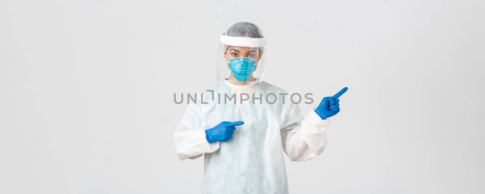 Covid-19, coronavirus disease, healthcare workers concept. Professional female doctor, tech lab employee in personal protective equipment pointing fingers right, showing way, white background by Benzoix