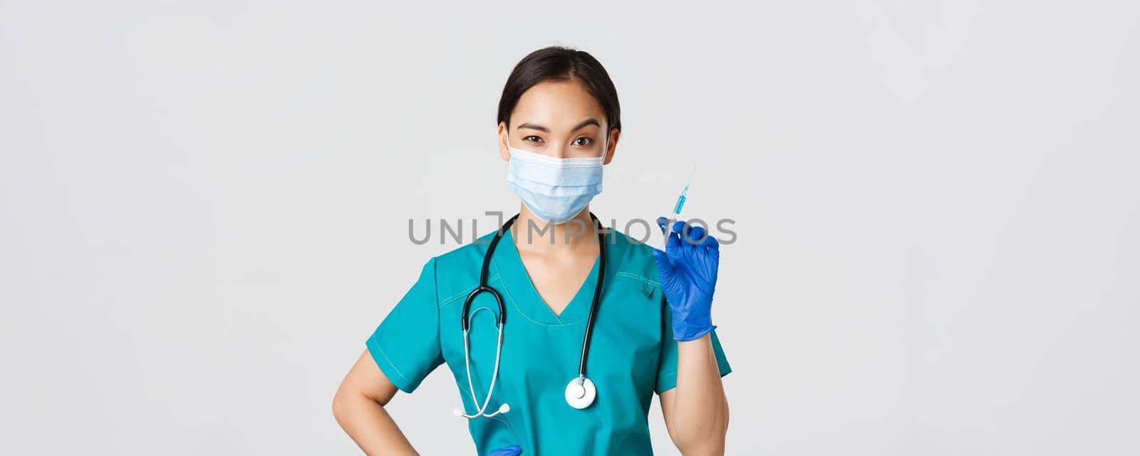 Covid-19, coronavirus disease, healthcare workers concept. Sassy cute asian female nurse, intern in medical mask and gloves holding syringe with vaccine, ready to make shot, white background by Benzoix