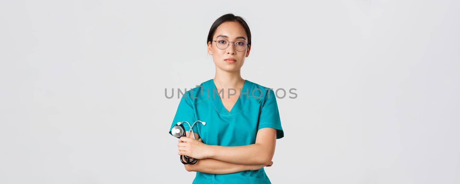 Covid-19, coronavirus disease, healthcare workers concept. Professional confident, serious-looking female doctor, physician in glasses and scrubs, holding stethoscope, standing white background by Benzoix