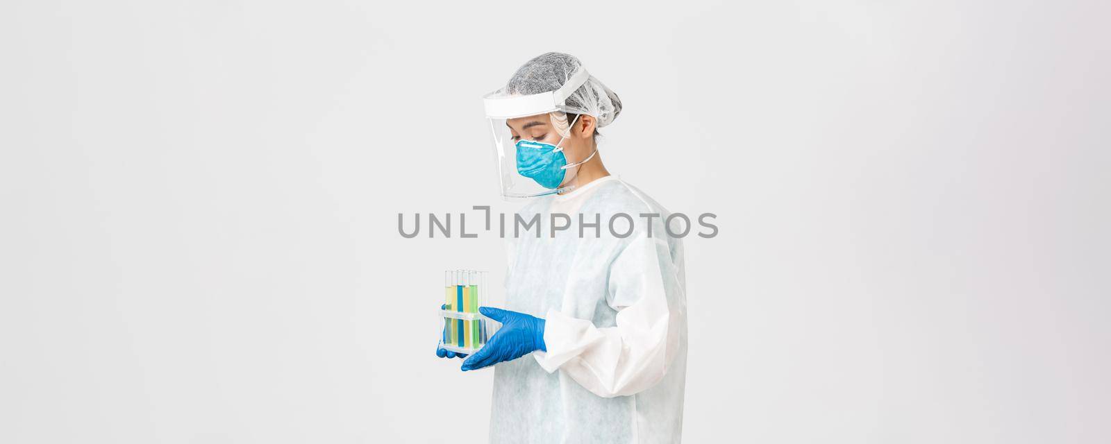 Covid-19, coronavirus disease, healthcare workers concept. Side view of careful serious asian female doctor, tech lab employee in personal protective equipment holding test-tube probes of vaccines.