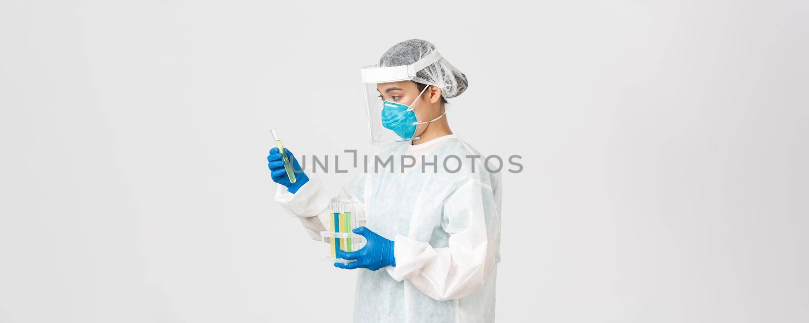 Covid-19, coronavirus disease, healthcare workers concept. Side view of careful serious asian female doctor, tech lab employee in personal protective equipment holding test-tube probes of vaccines.