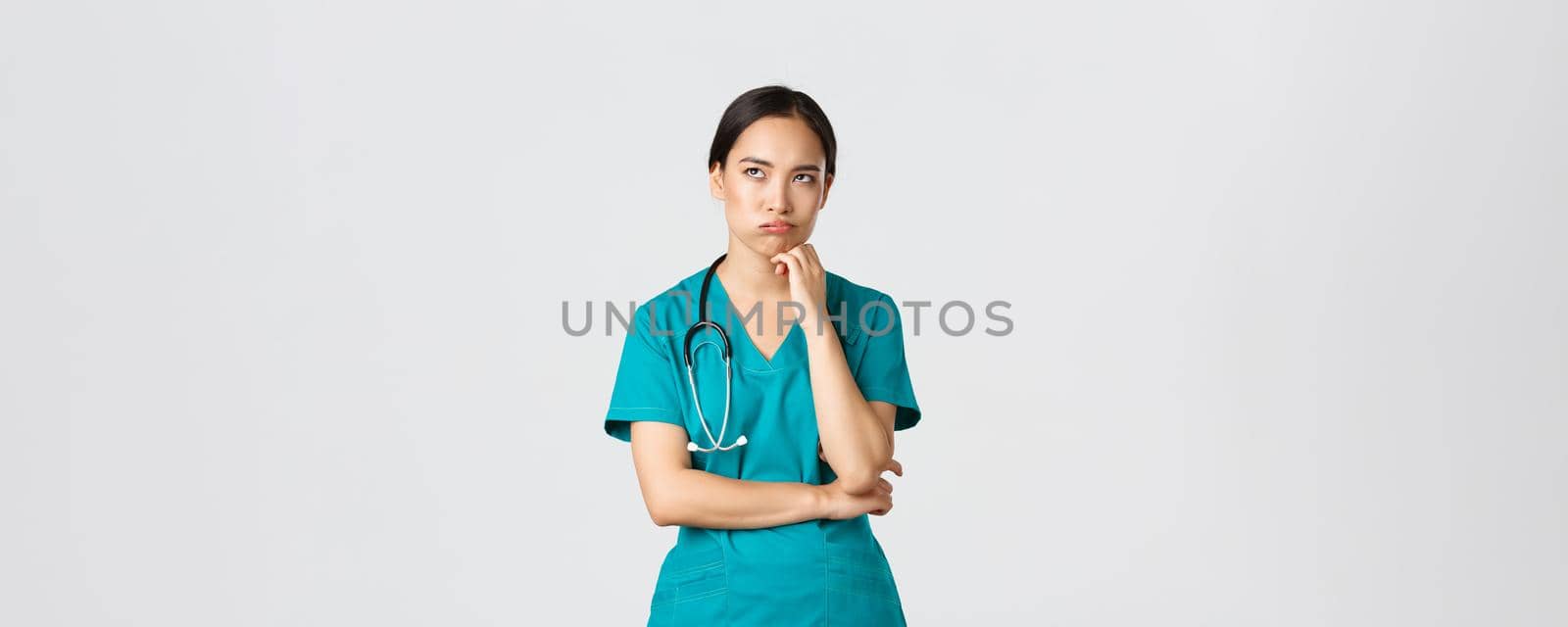 Covid-19, healthcare workers, pandemic concept. Annoyed and bothered asian female doctor, nurse on her shift looking irritated or tired, eye roll bored and exhale, standing white background in scrubs.