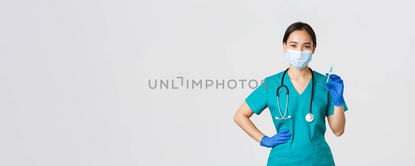 Covid-19, coronavirus disease, healthcare workers concept. Smiling confident asian female nurse in scrubs and medical mask, holding syringe with vaccine, doing vaccination shot, white background.