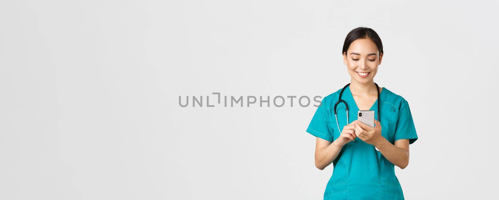 Covid-19, healthcare workers and preventing virus concept. Smiling happy beautiful asian female intern, doctor making phone call, looking at mobile phone screen pleased, messaging or using app by Benzoix