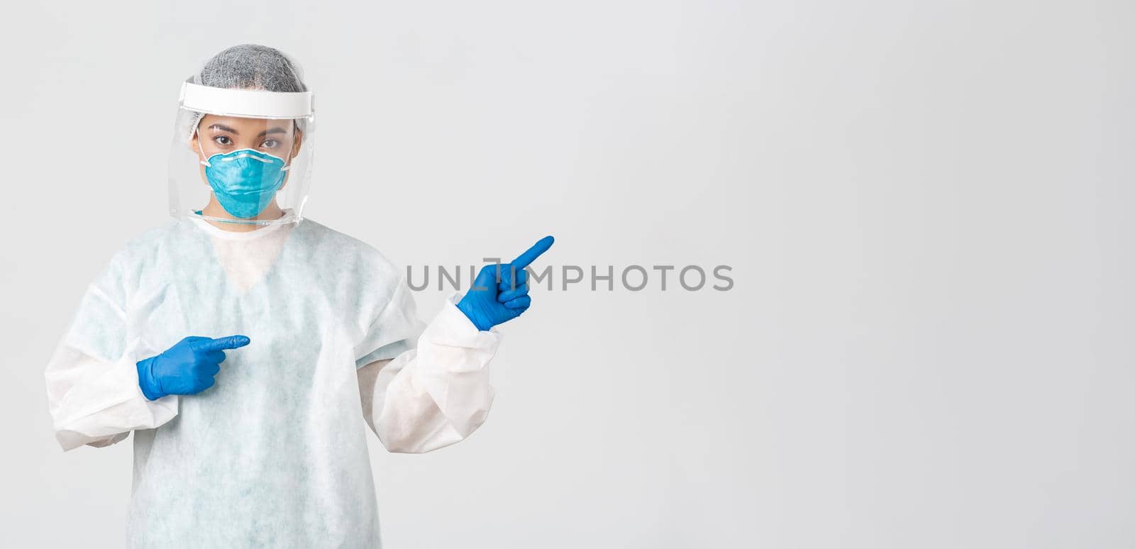 Covid-19, coronavirus disease, healthcare workers concept. Professional female doctor, tech lab employee in personal protective equipment pointing fingers right, showing way, white background.