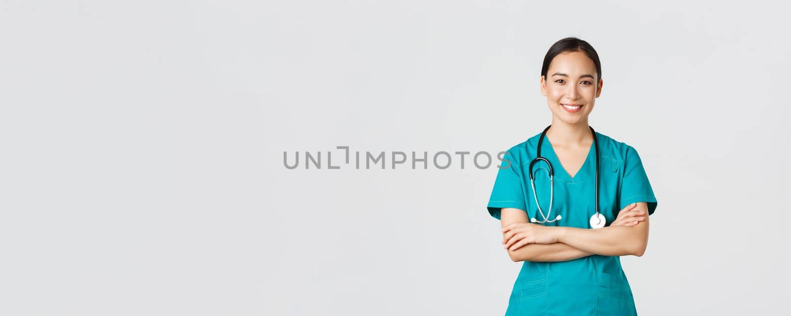 Covid-19, healthcare workers, pandemic concept. Portrait of confident smiling, attractive asian female nurse in scrubs, with stethoscope, cross arms chest and looking at camera, white background.