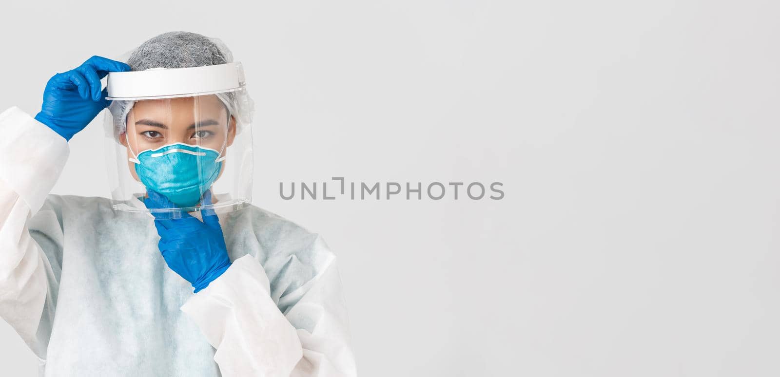 Covid-19, coronavirus disease, healthcare workers concept. Confident serious-looking female asian doctor, put on face shield and respirator, personal protective equipment, white background by Benzoix