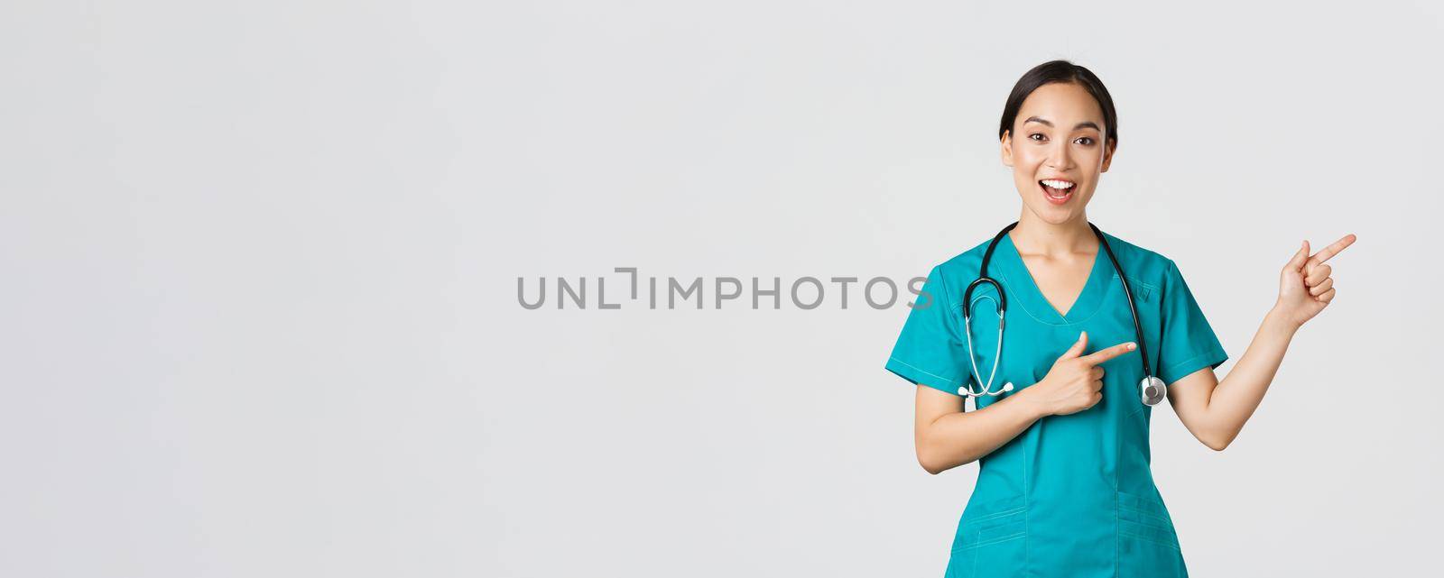 Covid-19, healthcare workers, pandemic concept. Glad smiling, beautiful asian doctor, female therapist in scrubs or nurse pointing fingers upper right corner, showing way to patient, white background by Benzoix