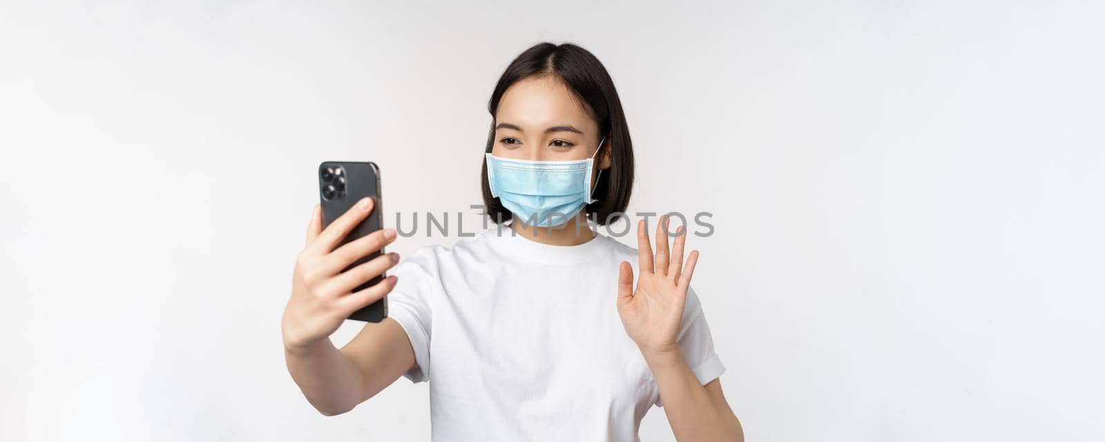 Health and covid-19 concept. Modern asian girl, student in medical mask, video chat with mobile phone, waving hand at smartphone app, standing over white background by Benzoix