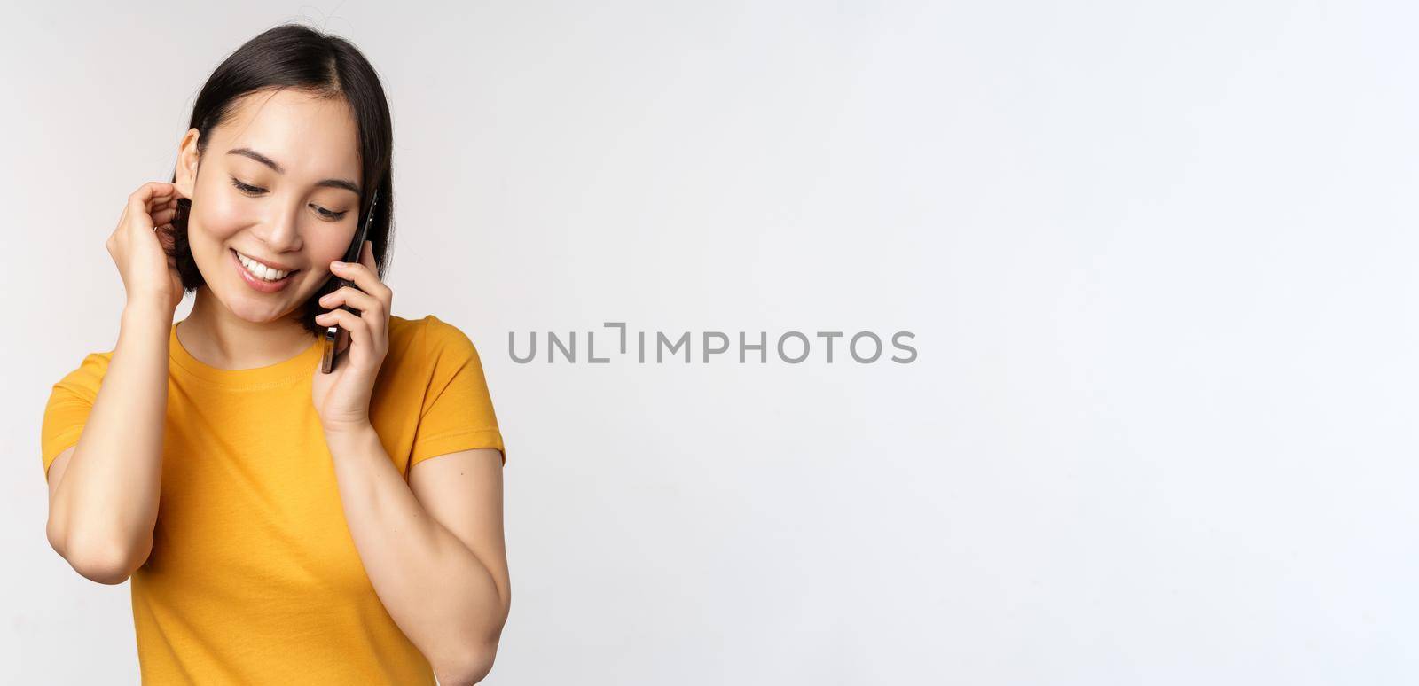 Romantic and cute asian girl answer phone call, talking on mobile, smiling coquettish, standing in yellow tshirt against white background.