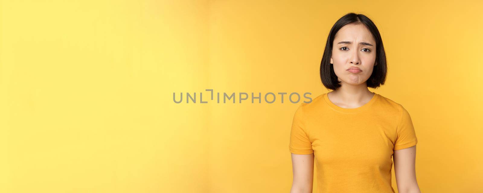 Disappointed asian girl sulking, looking upset, feel unair, standing in yellow t-shirt over white background by Benzoix