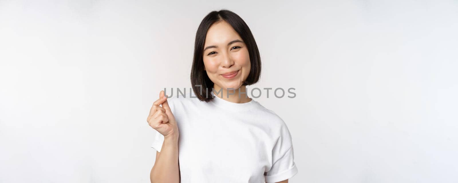 Beautiful asian woman smiling, showing finger hearts gesture, wearing tshirt, standing against white background.