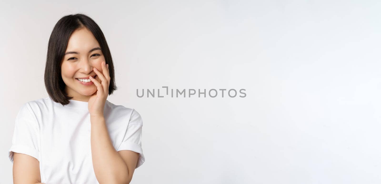 Portrait of young beautiful woman, korean girl laughing and smiling, looking coquettish, standing against white background by Benzoix