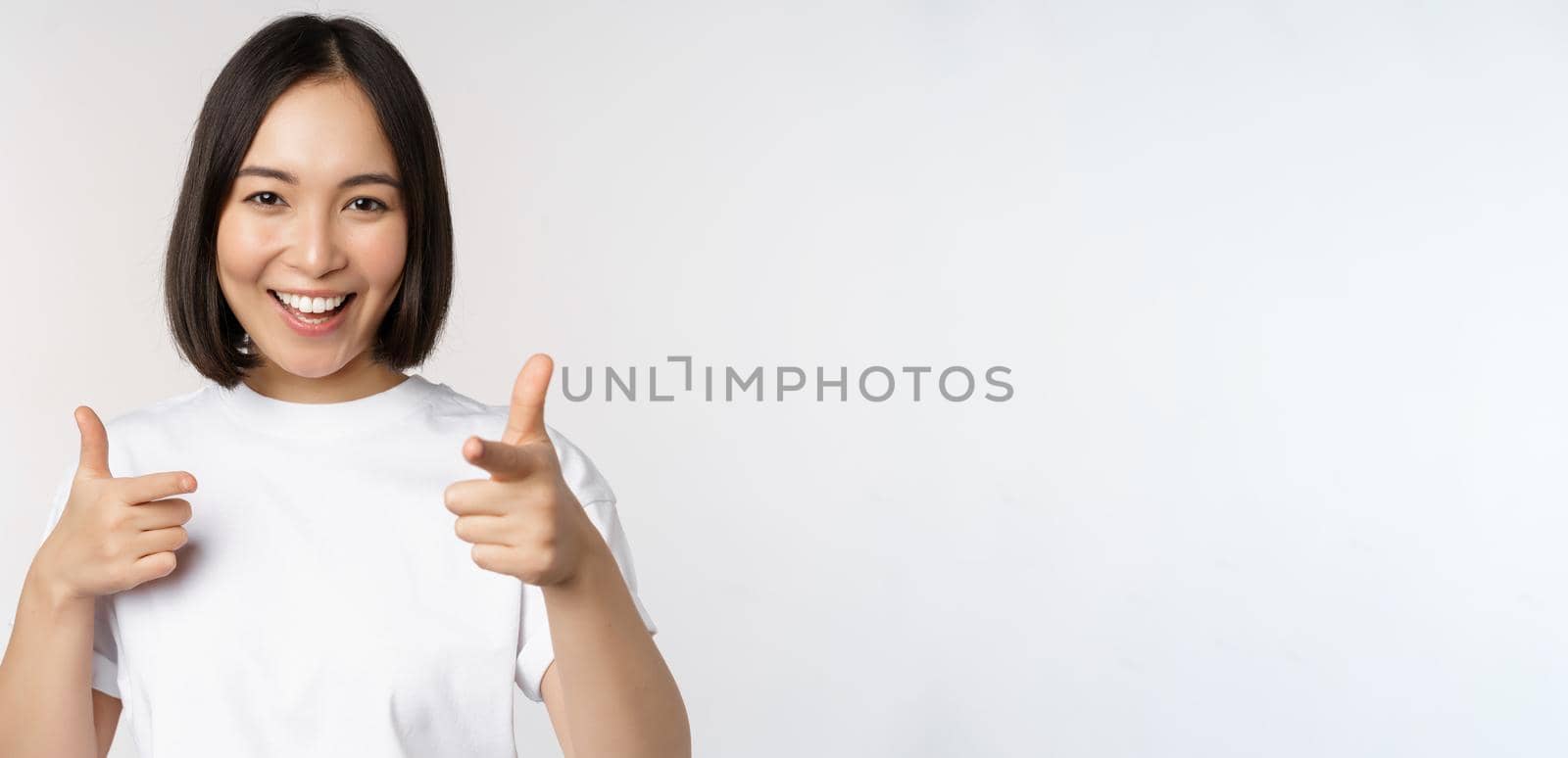 Image of smiling asian girl pointing fingers at camera, choosing, inviting you, congratulating, standing in tshirt over white background by Benzoix