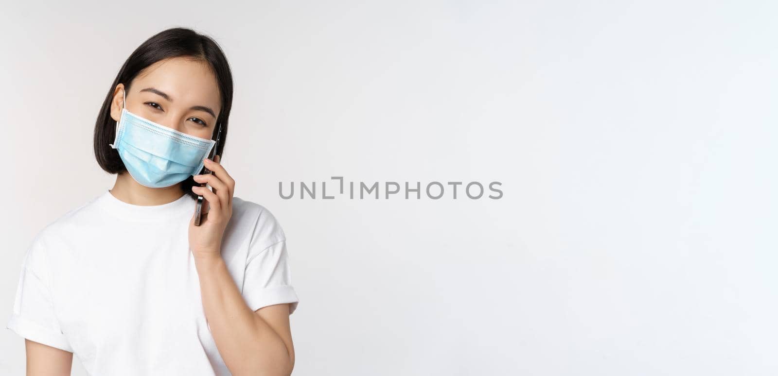 Health and covid-19 concept. Smiling asian girl in medical face mask talking on phone, answer mobile call, standing over white background.