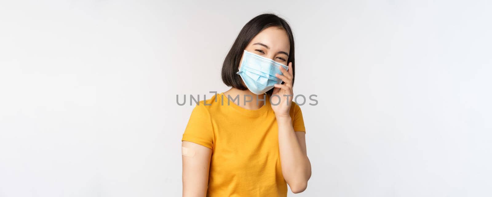 Covid-19, vaccination and healthcare concept. Cute asian girl in medical face mask, showing band aid after coronavirus vaccination, standing over white background.