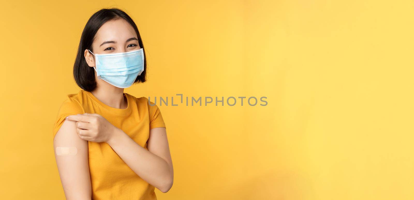 Vaccination and covid-19 pandemic concept. Smiling asian woman in medical face mask, showing her shoulder with band aid after vaccinating from coronavirus, yellow background by Benzoix